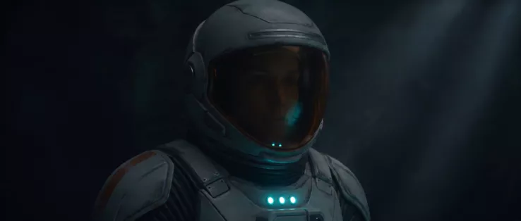 Close on the astronaut in her helmet in a dark interior, a shaft of light from above, three cyan indicators lit at her collar