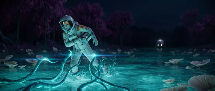 The astronaut bent forward in the shallows at night, glowing tendrils wrapped around her legs, magenta forest and the lander’s lights behind her